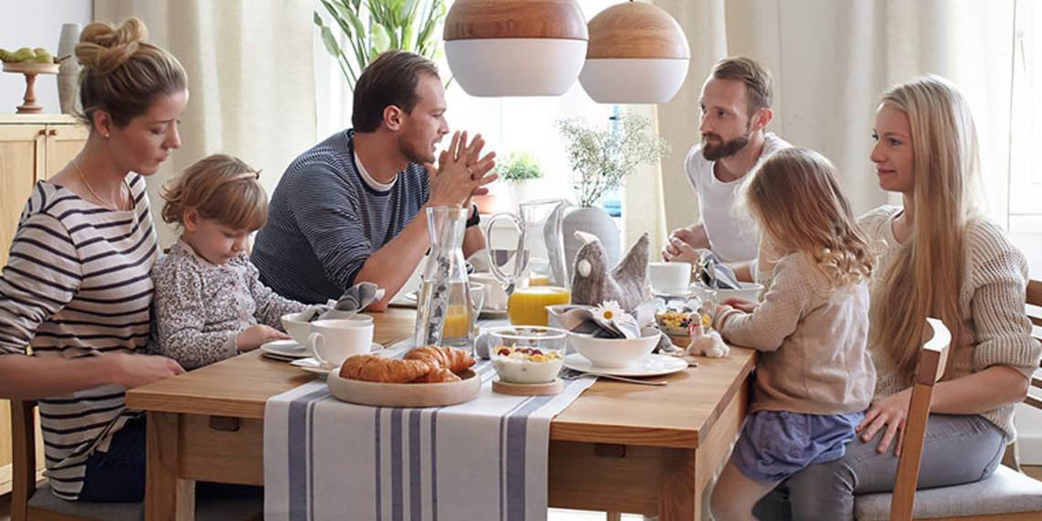 Zwei junge Familien mit Kindern sitzen an einem Frühstückstisch.